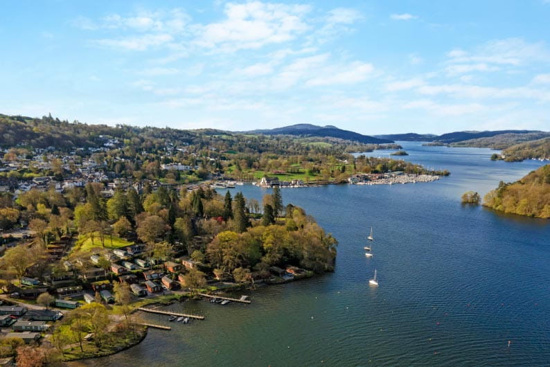 An aerial view over Lake Windermere in the Lake District, with caravans and lodges at Fallbarrow Holiday Park visible along the shore