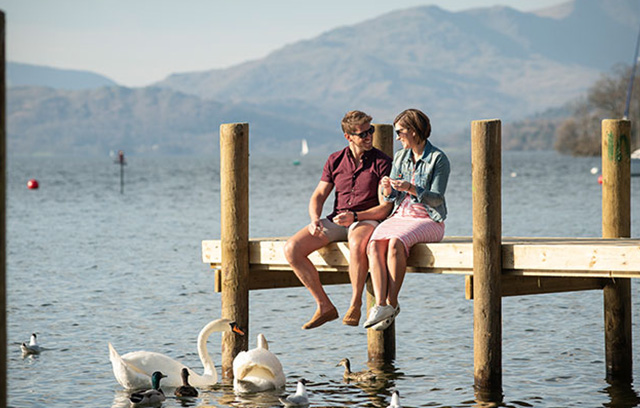A couple sitting on a jetty at Lake Windermere with swans in the water