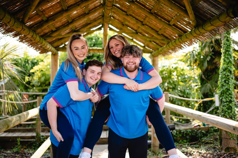 Four people wearing matching blue uniforms pose happily under a wooden gazebo. Two of them are giving piggyback rides, smiling and enjoying the moment.