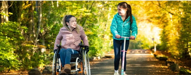 a woman in a wheelchair riding along a lane with a girl on a scooter