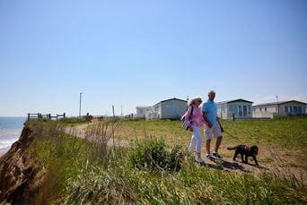 A couple walking a dog along a coastal path, with caravans at a Parkdean Resorts holiday park in the background