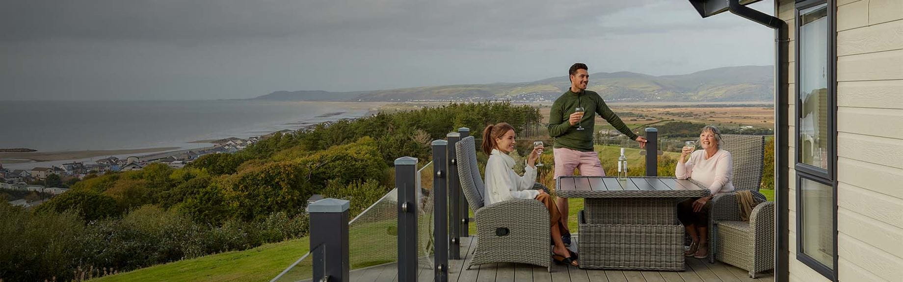 A family enjoying drinks on the veranda of their holiday lodge, overlooking the Welsh coast