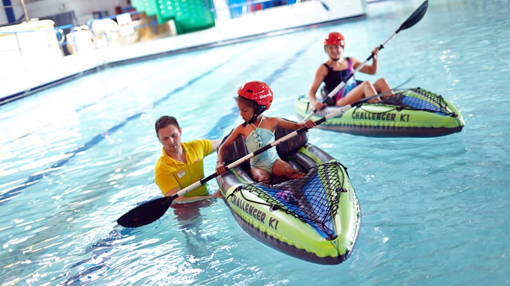 Kids and a lifeguard in an indoor swimming pool trying kayaking