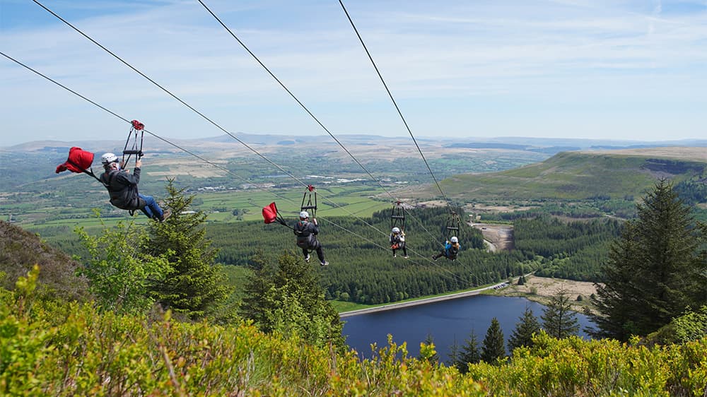 People zooming down a very long zip line over a reservoir in the Welsh countryside
