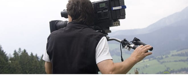 A man holding filming equipment overlooking trees and mountains