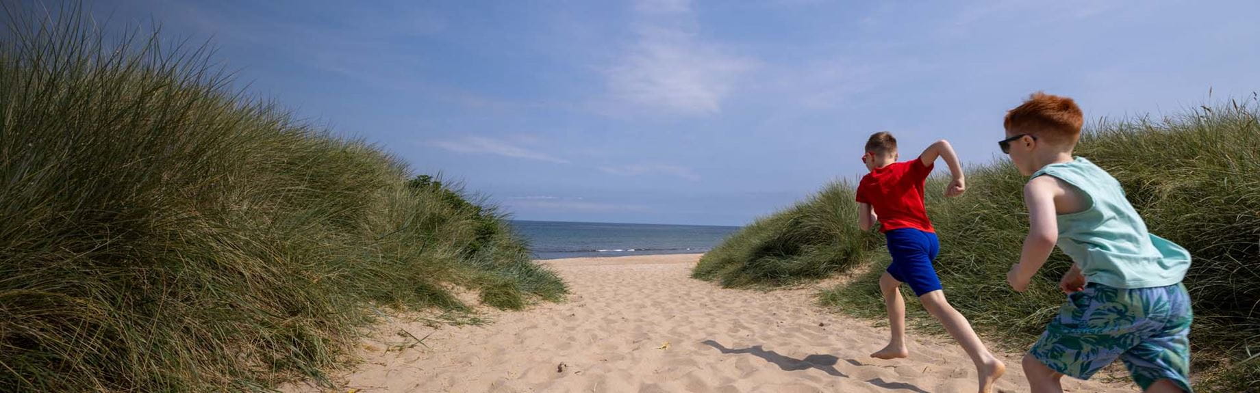 Two boys running along the sand between the dunes towards the beach
