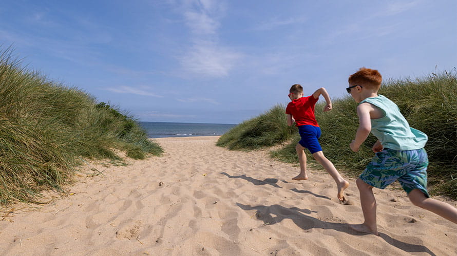 Two boys running along the sand between the dunes towards the beach