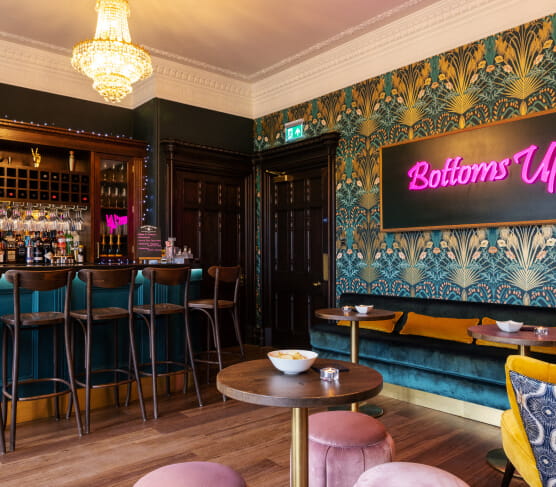 Bar with high bar stool, funky patterned blue wallpaper with pink neon 'Bottoms Up' sign
