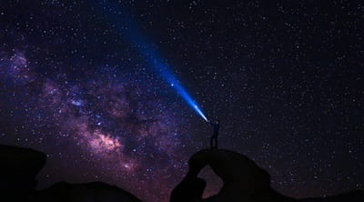 A man standing on top of a rock shining a torch into a dark, starry sky