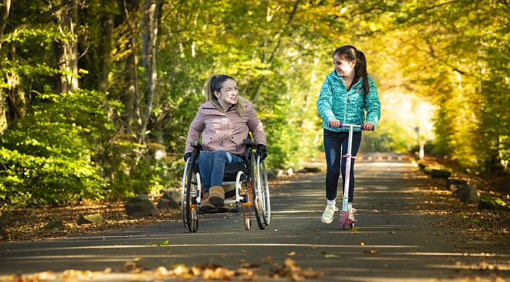a woman in a wheelchair riding along a lane with a girl on a scooter