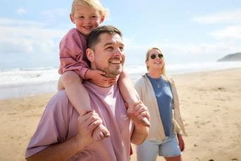 a family cene on the beach with child on a mans shoulder with a woman in the background