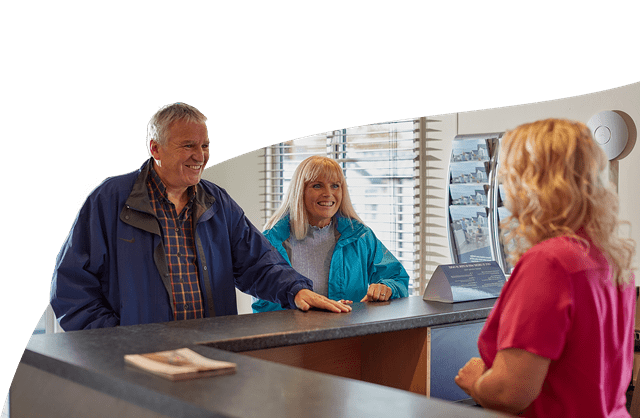 Couple smiling stood at a reception desk speaking with a member of staff at Parkdean Resorts