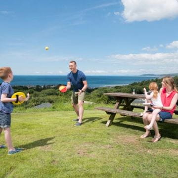 Man and boy playing catch in a park while a woman and girl sit watching on a wooden bench