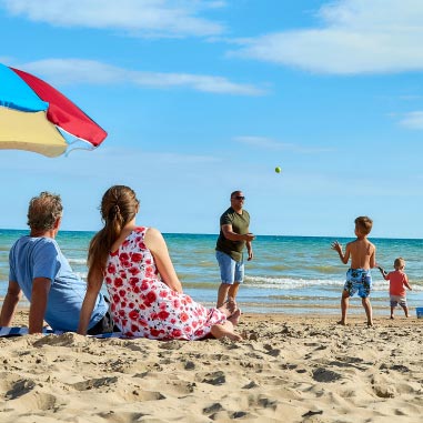 Family playing catch on the beach with a couple sat under a sun umbrella watching