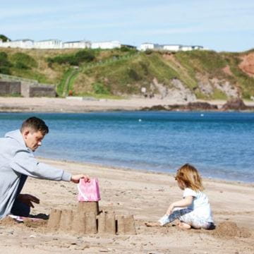 Father and child building a sandcastle on a beach with a clifftop coastal town in the background