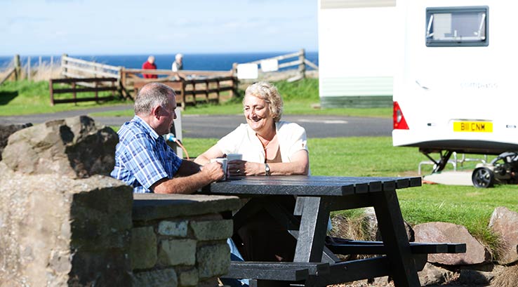 A couple enjoying coffees on a picnic bench in the sun by their touring caravan