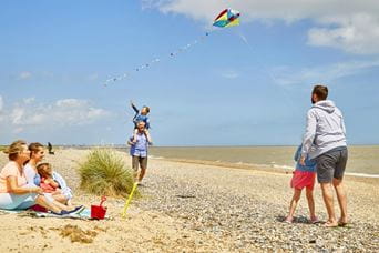 A man and two kids flying a kite on a beach with two women and a baby sat watching on