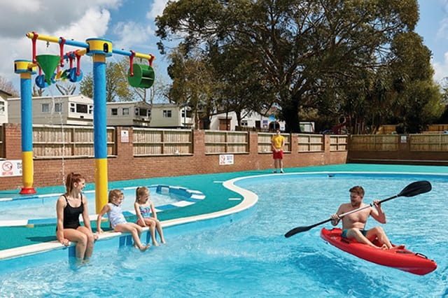 A man kayaking in the outdoor swimming pool whilst his family watches from the side