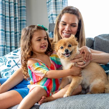 Mother and daughter sitting on a sofa petting a small tan dog