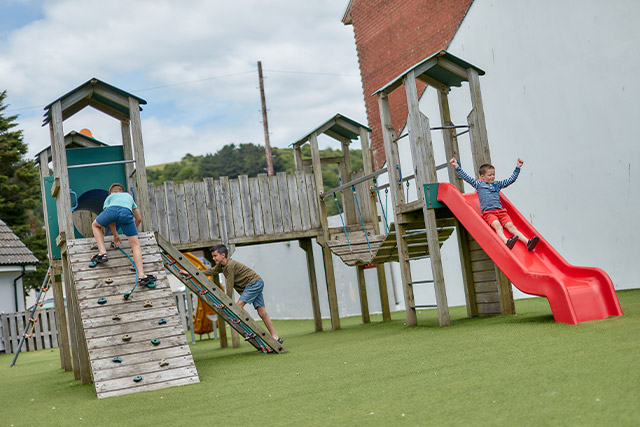 A boy sliding down the slide on the adventure playground whilst his mother supervises