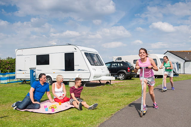 A family enjoying a picnic outside their touring caravan and a girl riding her scooter at Southerness Holiday Park