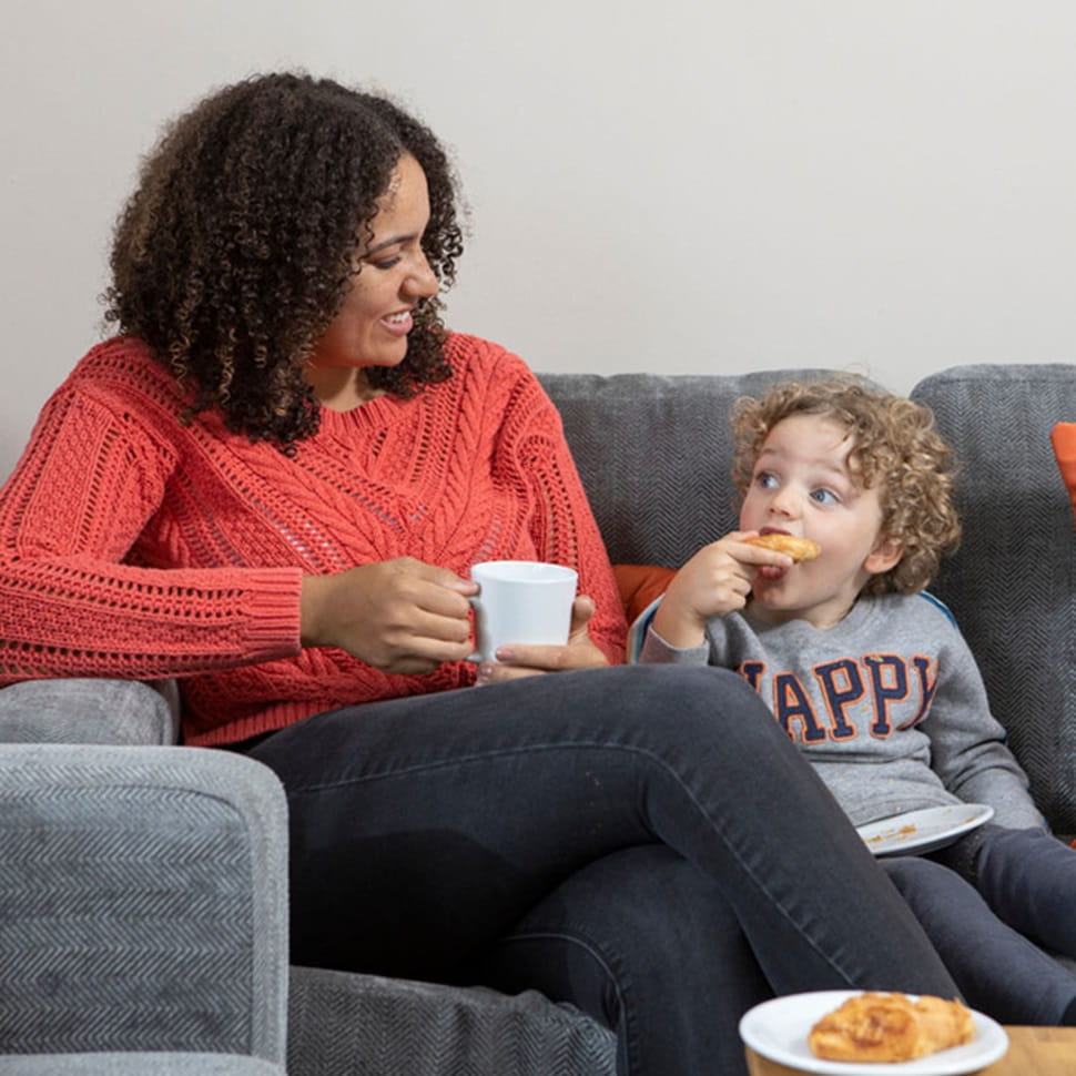 A mother and little boy sitting on a sofa with drinks and snacks