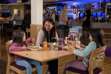 Family enjoying a meal in a restaurant with couple leaning on a bar counter in the background