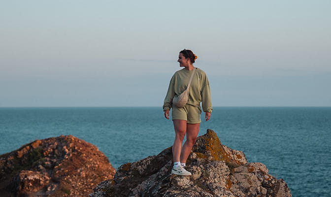 A girl standing on a rocky headland looking at the views of the Cornish coast at sunset