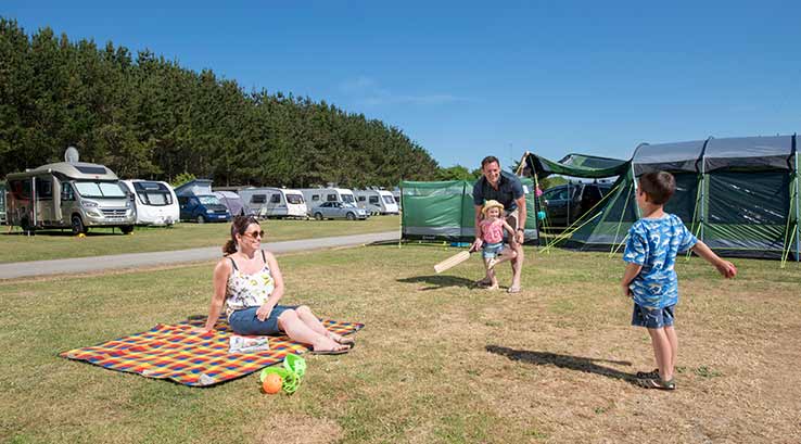 Family playing cricket next to a tent in camping field on a sunny day