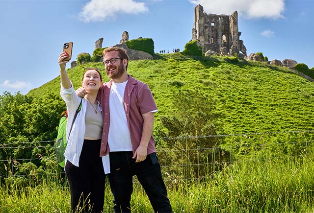 A couple taking a selfie in front of Corfe Castle in Dorset on a sunny day