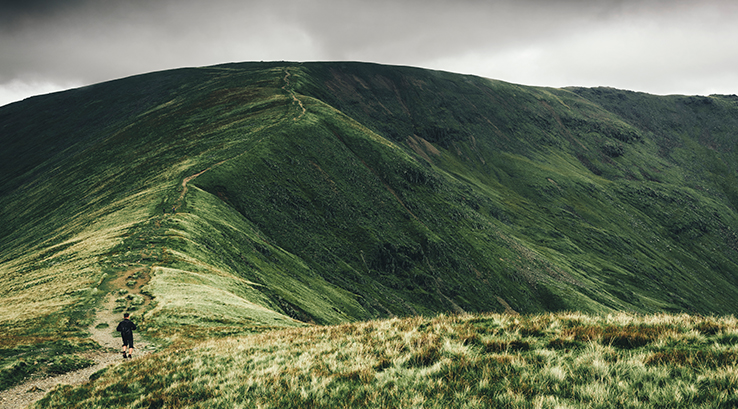 A man walking across the grassy mountain peaks along the Fairfield Horseshoe in the Lake District