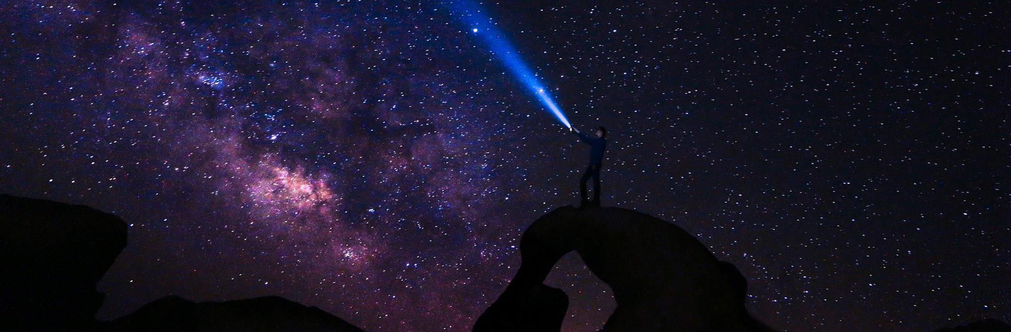 A man standing on top of a rock shining a torch into a dark, starry sky