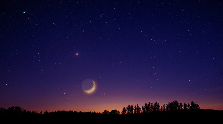 A starry sky and moon with a sun setting over the silhouette of a landscape