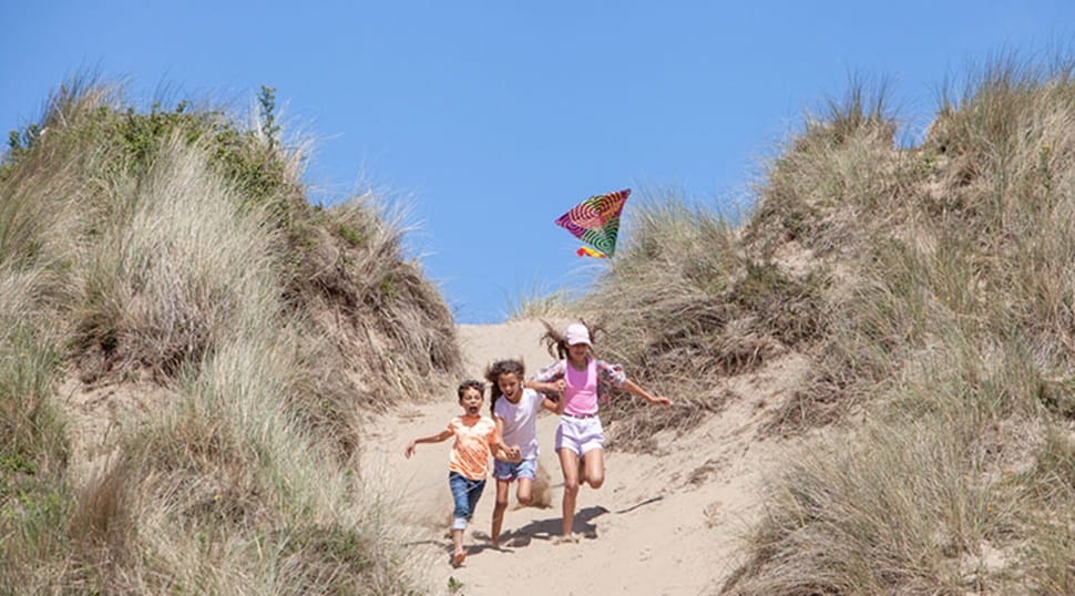 three children running down a sand dune with a kit