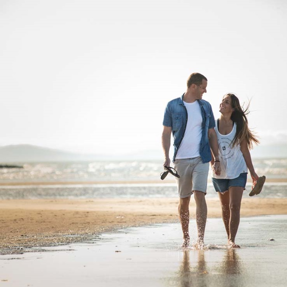 A couple walking barefoot along the shoreline of a beach in Wales