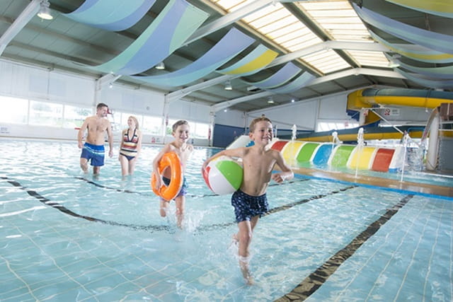 A family walking across the indoor swimming pool with beach balls and rubber rings